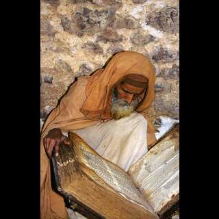 A spiritual man in robes, brown skin, grey beard, reading from a massive tome.